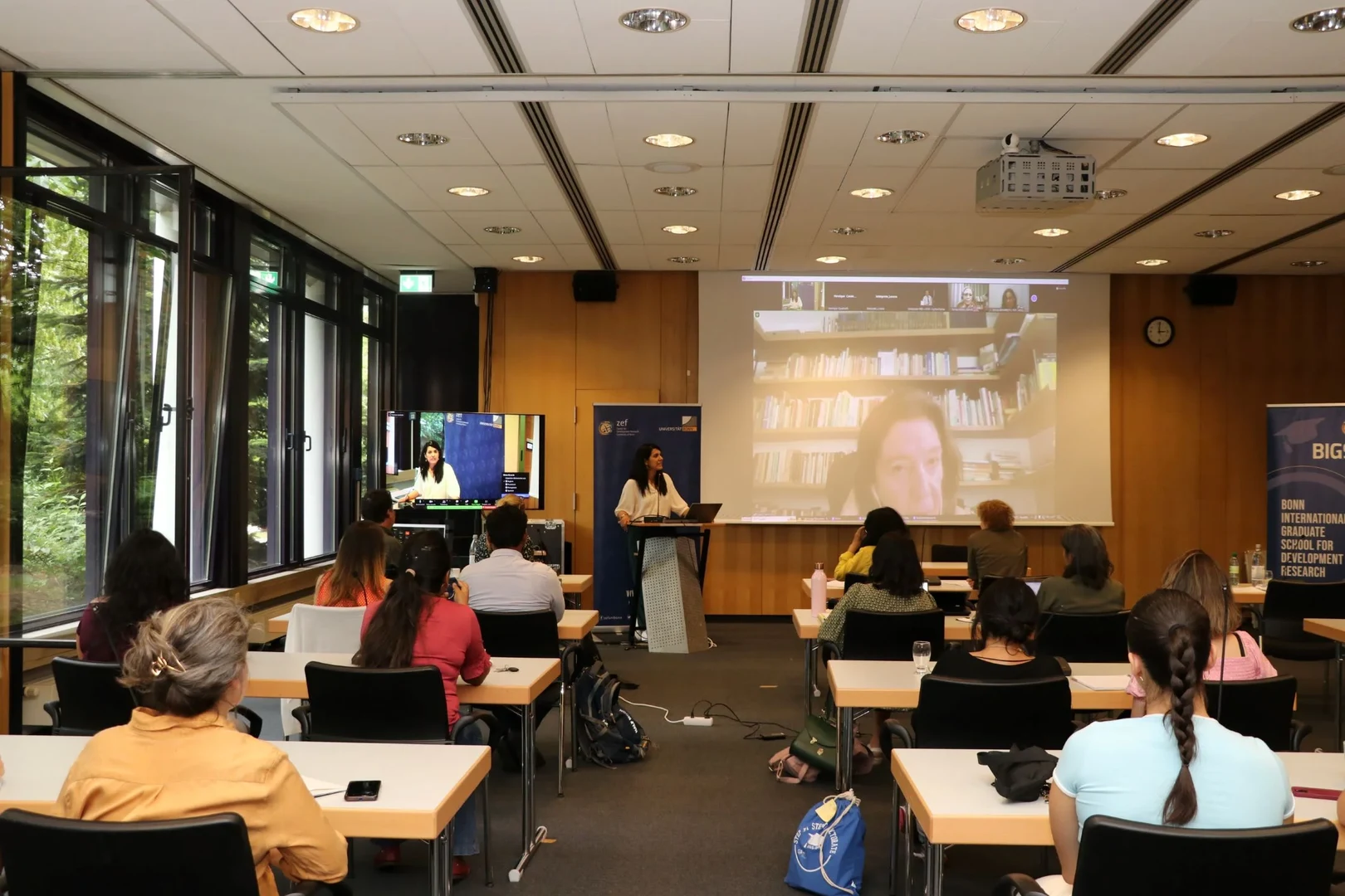 Eine Wissenschaftlerin und ein Wissenschaftler arbeiten hinter einer Glasfassade und mischen Chemikalien mit Großgeräten.